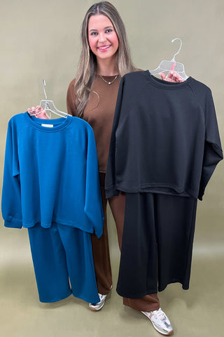 Woman standing between two outfits on hangers, one blue and one black, against a plain background.