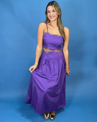 A woman modeling a purple two-piece crop top and maxi skirt set with silver rhinestone trim, standing against a solid blue studio backdrop.