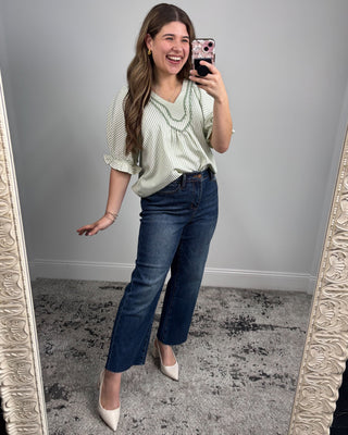 Woman taking a mirror selfie wearing a green top and blue jeans.