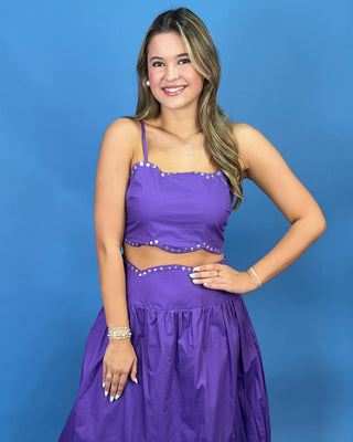 A woman modeling a purple two-piece maxi dress with a crop top and flared skirt, featuring silver rhinestone trim, set against a solid blue studio background.