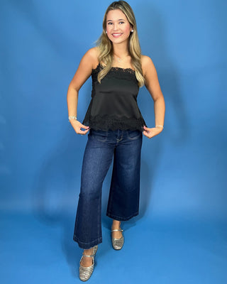A woman modeling a black satin strapless top with lace trim and dark blue wide-leg jeans