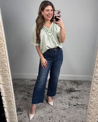 Woman taking a mirror selfie wearing a green striped blouse and blue jeans.