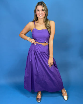 A woman modeling a purple two-piece maxi dress with a crop top and flared skirt, featuring silver rhinestone trim, set against a solid blue studio background.