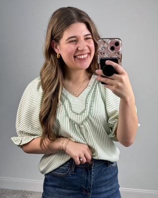 Woman taking a mirror selfie wearing a green striped shirt and blue jeans on a gray background