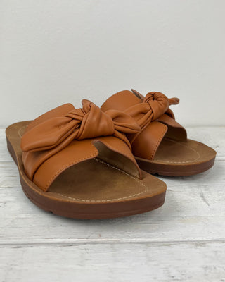 Pair of brown sandals with bow detail on a light wooden floor.