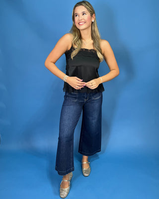 A woman modeling a black satin strapless top with lace trim and dark blue wide-leg jeans