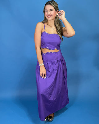A woman modeling a purple two-piece maxi dress with a crop top and flared skirt, decorated with silver rhinestone trim, against a solid blue studio background.