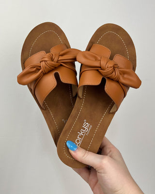 Brown sandals with bow details held by a hand against a light gray background