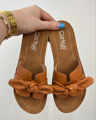 Brown sandals with bow detail held by a hand against a white background
