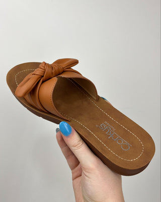 Brown sandal with a knot detail held by a hand against a light gray background