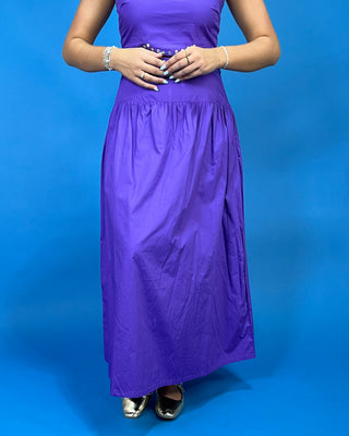 A woman wearing a sleeveless purple maxi dress with a gathered waist and silver flats, standing against a solid blue background.