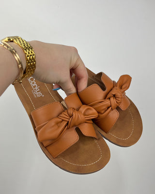 Brown sandals with bow details held by a hand against a plain background