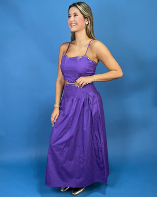 A woman modeling a purple spaghetti-strap maxi dress with silver rhinestone trim at the bust and waist, standing against a solid blue studio backdrop.