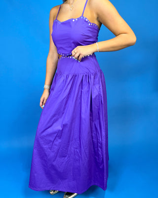 A woman modeling a bright purple maxi dress with thin straps, a beaded waistband, and a gathered skirt, set against a solid blue studio background.