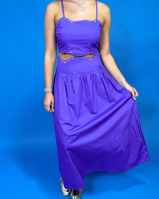 A woman modeling a bright purple two-piece maxi dress with a crop top and flared skirt, featuring silver rhinestone trim along the neckline and waist, set against a solid blue studio background.