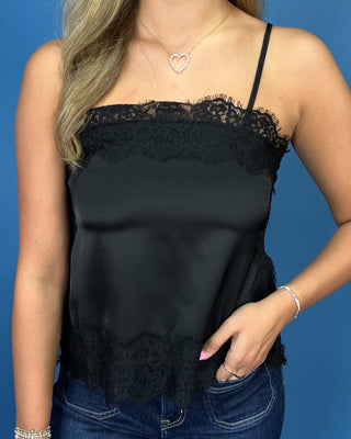 A woman wearing a black satin camisole top with black lace trim at the neckline and hem, paired with blue jeans