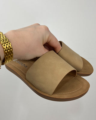 Beige sandal held by a hand with a gold bracelet on a light gray background