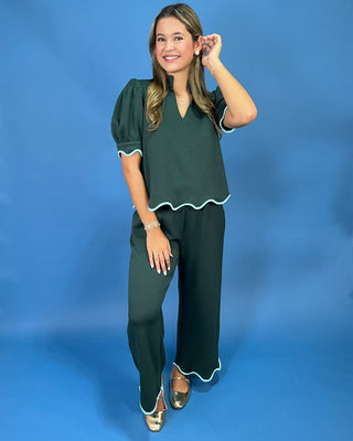 A woman modeling a dark green two-piece outfit consisting of a short-sleeve top with puff sleeves and a scalloped hem, paired with matching wide-leg pants with white piping trim, set against a solid blue studio background.