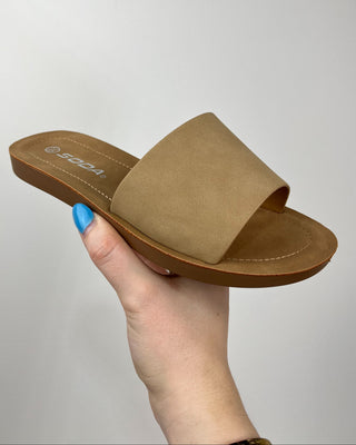 Brown sandal held by a hand with blue nail polish against a gray background