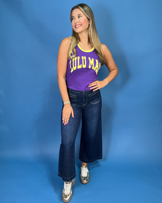 A woman modeling a purple sleeveless tank top with yellow trim and 'LULU MAC' printed in yellow across the front, paired with dark blue jeans