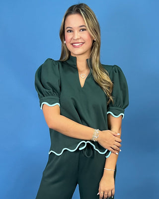 A woman modeling a dark green puff-sleeve top with light blue scalloped trim, paired with matching dark green pants, set against a solid blue studio background.