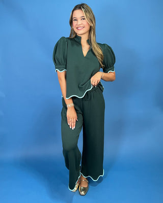 A woman modeling a dark green two-piece jumpsuit with light green piped trim, featuring puff sleeves and wide-leg pants, set against a solid blue studio background.