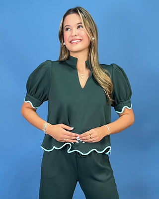 A woman modeling a dark green puff-sleeve top and matching pants set against a solid blue studio background. The outfit features light blue ruffle trim along the collar, sleeve cuffs, and hem.