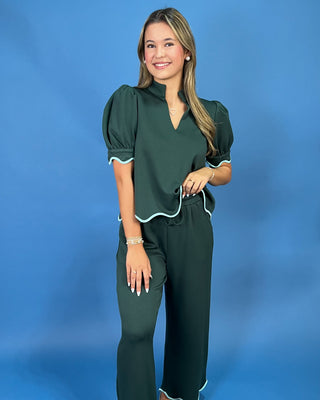 A woman modeling a dark green two-piece jumpsuit with white piping trim, featuring puff sleeves and wide-leg pants, set against a solid blue studio background.