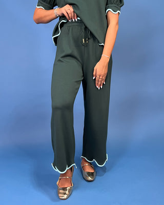 A model wearing dark green wide-leg pants with light blue scalloped trim and a matching dark green top, standing against a solid blue studio background.