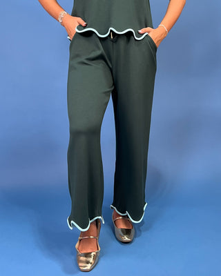 A person wearing blue cropped wide-leg jeans with a beige top and metallic sandals, standing against a solid pink studio background.