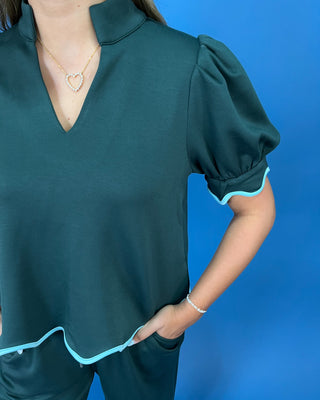 A woman wearing a dark teal green short-sleeve top with puff sleeves and light blue trim, paired with matching dark green bottoms, set against a solid blue background.