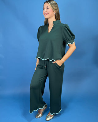 A woman modeling a dark green two-piece set consisting of a short-sleeve blouse with puff sleeves and scalloped trim, and matching wide-leg pants with scalloped hems, set against a solid blue studio background.
