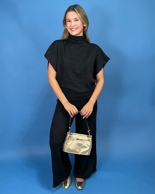 A woman modeling a black short-sleeve mock-neck top and black wide-leg pants, holding a metallic gold handbag against a solid blue studio background.