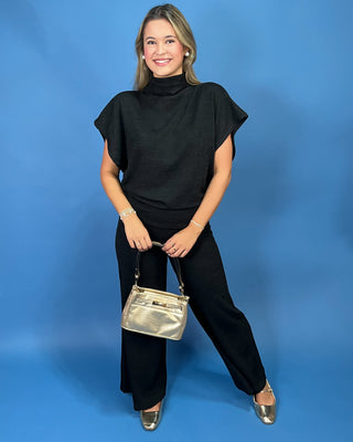 A woman modeling a black turtleneck top with short flutter sleeves and black wide-leg pants, holding a metallic silver handbag against a solid blue studio background.