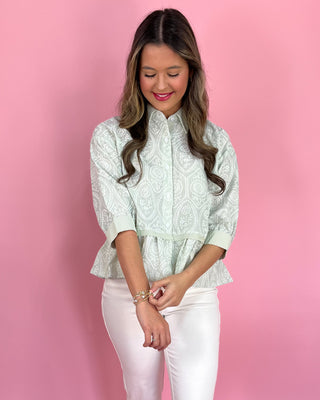Woman wearing a light green patterned blouse and white pants against a pink background