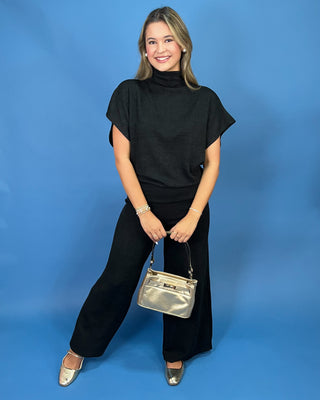 A woman posing in a black short-sleeve mock neck top and black wide-leg pants, holding a metallic silver handbag against a solid blue background.