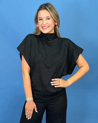 A woman modeling a black short-sleeve mock neck top with draped cap sleeves, paired with black pants, standing against a solid blue studio background.