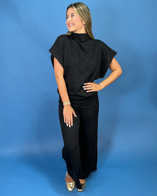 A woman modeling a black high-neck, short-sleeved maxi dress with draped cap sleeves, standing against a solid blue studio backdrop.