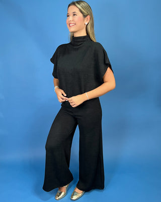 A woman modeling a black two-piece outfit consisting of a short-sleeve mock-neck top and matching wide-leg pants, standing against a solid blue studio backdrop.
