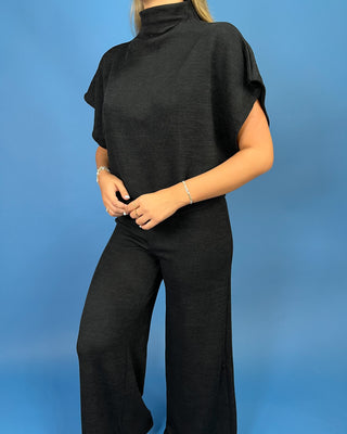 A woman wearing a black mock-neck, short-sleeved draped top and matching wide-leg trousers, standing against a solid blue studio background.