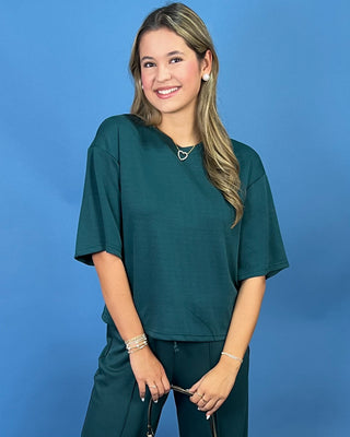 A woman wearing a dark teal green short-sleeve scrub top and matching scrub pants, standing against a solid blue studio background.