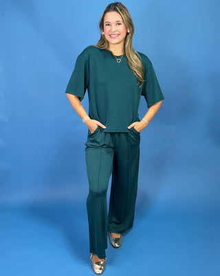 A woman modeling a matching dark green two-piece set consisting of a short-sleeve top and wide-leg pants, standing against a solid blue studio backdrop.