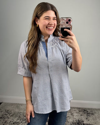 Woman taking a mirror selfie wearing a blue and white striped shirt against a gray wall.