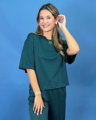 A woman modeling a dark teal green scrub top and matching scrub pants against a solid blue studio background.