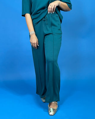 A person wearing a matching teal green lounge set consisting of a short-sleeve top and wide-leg drawstring pants, standing against a solid blue studio background.