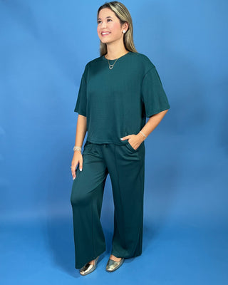 A woman modeling a dark teal green two-piece lounge set consisting of a short-sleeve top and matching wide-leg pants, standing against a solid blue studio backdrop.