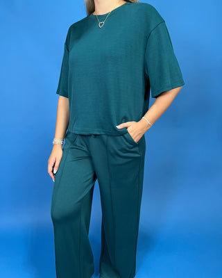 A woman wearing a matching dark teal green two-piece lounge set consisting of a short-sleeve crewneck top and wide-leg pants, posed against a solid blue studio background.