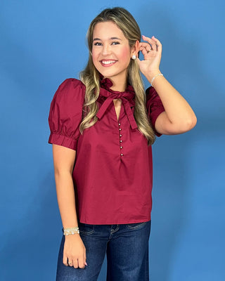 A woman modeling a short-sleeve burgundy blouse with puff sleeves, a keyhole tie neckline, and front buttons, paired with blue jeans