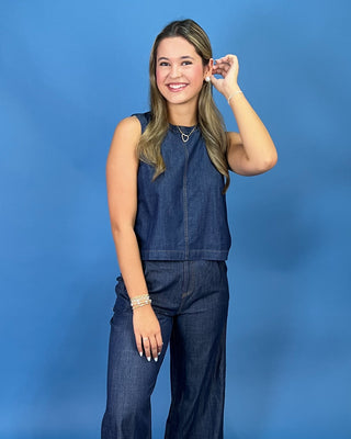 A young woman with blonde hair stands against a solid blue studio background, wearing a sleeveless dark blue denim top and matching wide-leg denim pants.