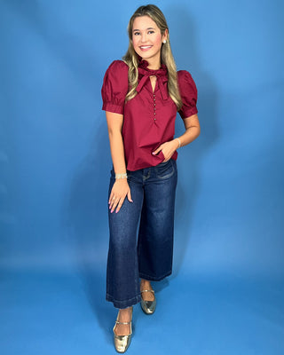 A woman modeling a short-sleeve burgundy blouse with puff sleeves, a keyhole tie neckline, and dark blue wide-leg jeans